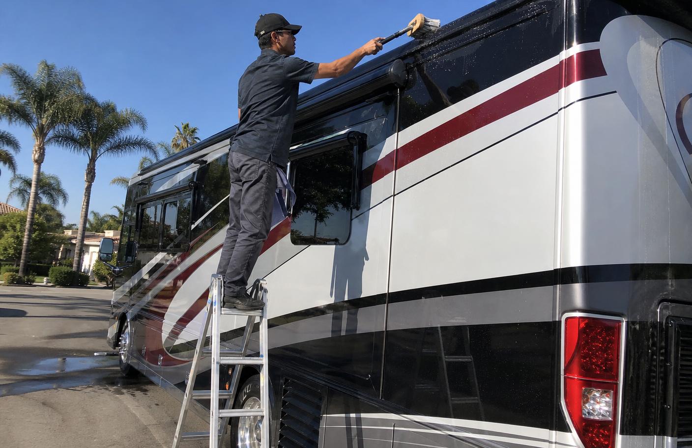 Detailer on a step ladder cleaning the upper sidewall and roof edge of a motorhome